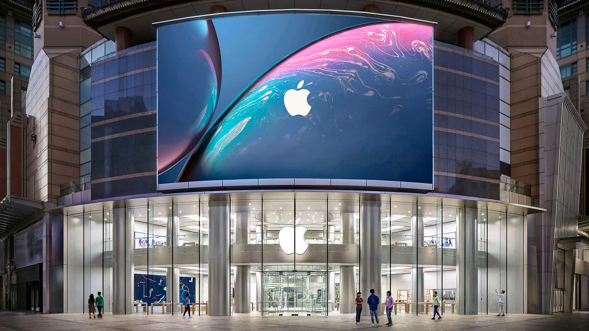 Exterior of Apple Wangfujing, located in the center of Beijing. The curved glass storefront takes up a corner of the mall, spans 2 stories high and the store has a fan-shaped layout. There is also a large LED screen that sits on top of the store.